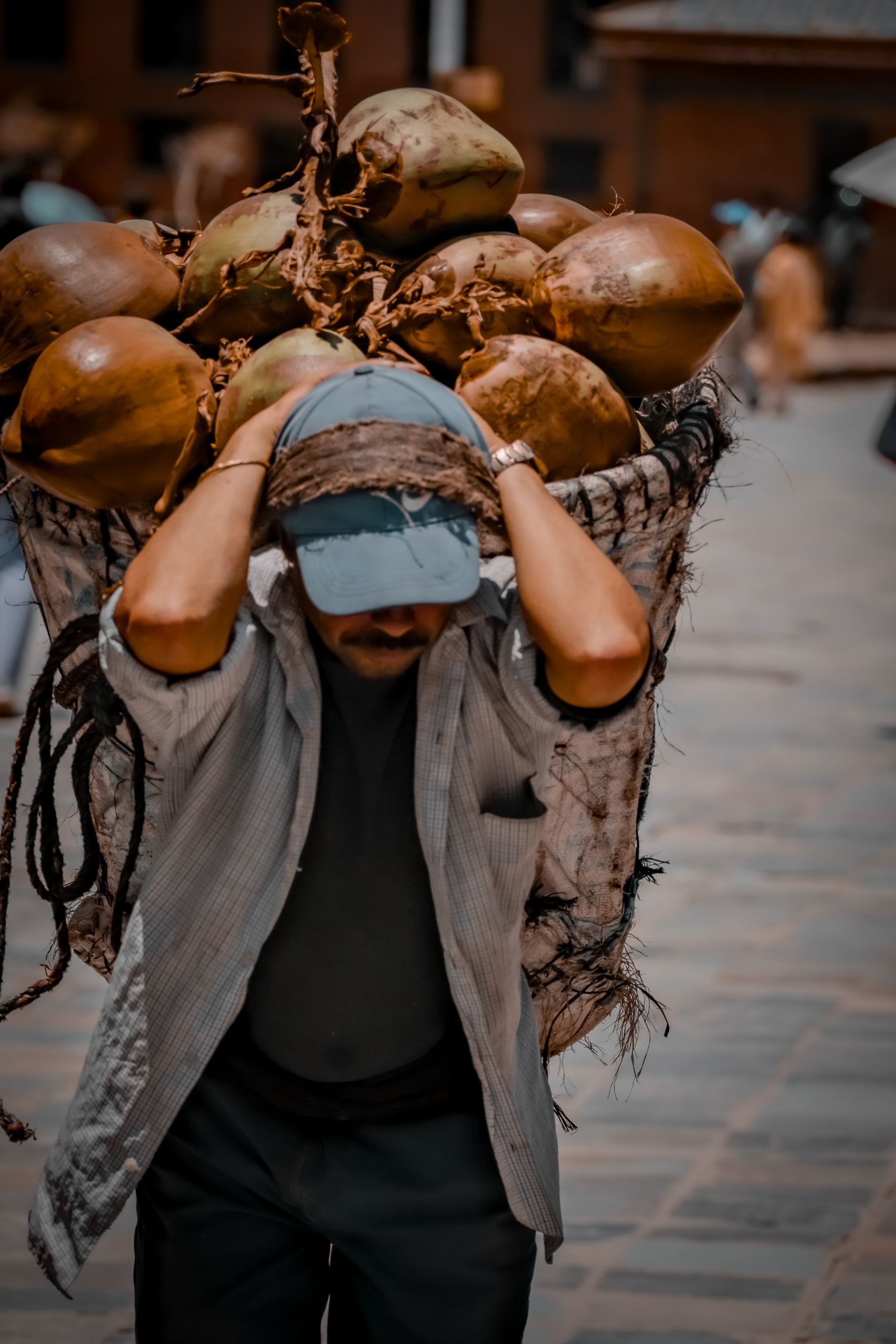 A man struggles to carry a load of coconuts in a bustling street.